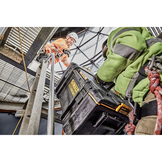 A worker in safety gear is carrying a DEWALT DWST83294-1 ToughSystem storage case up a metal staircase, with another worker above assisting. The scene is set in an industrial environment.