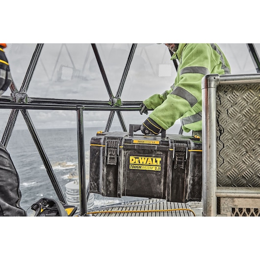 A person wearing a green high-visibility jacket is holding a DEWALT ToughSystem 2.0 tool box. The scene takes place inside an industrial structure near the ocean, with metal railings and steps visible.