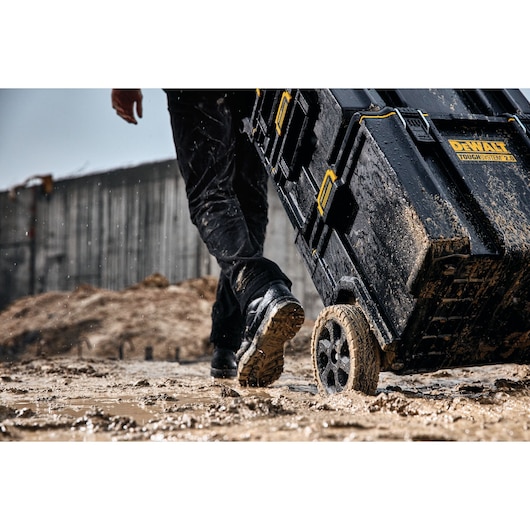 Close-up of a person wheeling a muddy DEWALT ToughSystem 2.0 tool storage box on a construction site, with wet ground and splashes of mud.
