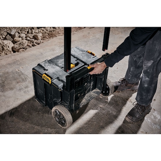 A person is pulling a DEWALT rolling tool storage box with large wheels and a telescopic handle on a dusty construction site.
