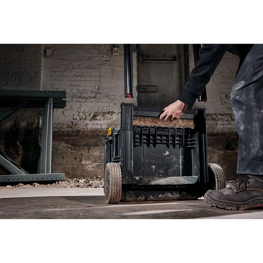 A DEWALT rolling tool box (model DWST83295-1) is shown being pulled by a person in a work environment. The box is black, has large rugged wheels, and a sturdy handle, suitable for carrying tools and materials.