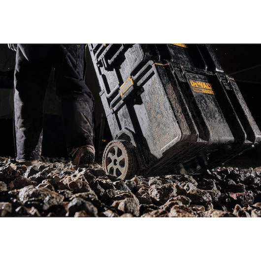Close-up view of a DEWALT ToughSystem rolling toolbox being wheeled over rough, rocky ground by a person. The toolbox and wheels appear covered in mud, highlighting its rugged durability for use in tough environments.