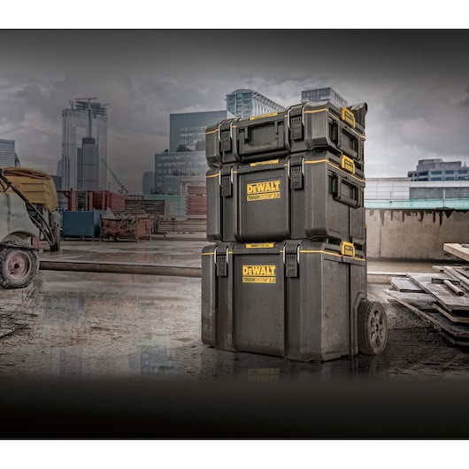 Three stacked DEWALT ToughSystem 2.0 tool storage boxes are shown on a construction site. The boxes are black with yellow accents and have wheels for easy transport. Modern city buildings are visible in the background.
