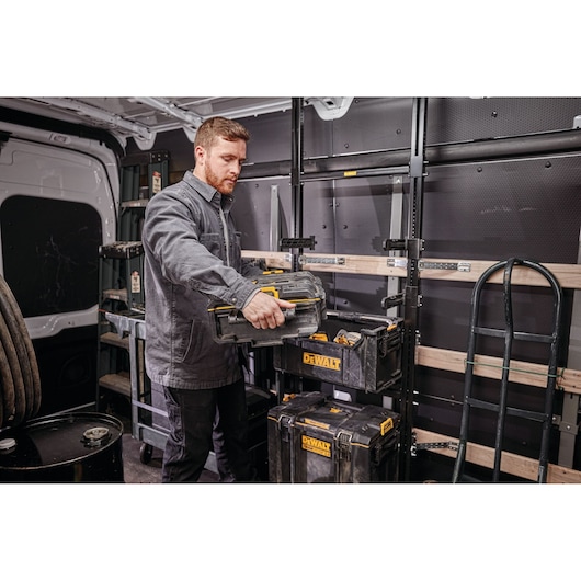 A person in work attire is organizing DEWALT toolboxes inside a van equipped with shelves and storage racks. The individual's face is blurred for privacy.