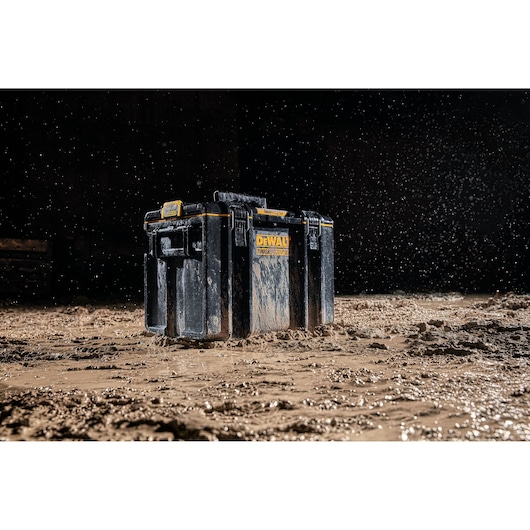 A black DEWALT tool box with yellow accents stands on muddy ground with water droplets in the air, showcasing its durability and rugged design for use in tough environments.