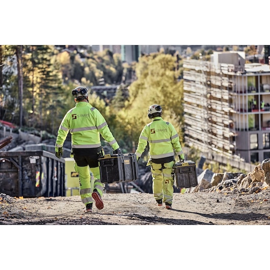 Two construction workers in high-visibility jackets and helmets walk on a construction site, each carrying a DEWALT tool box. The site is outdoors with buildings and trees in the background.