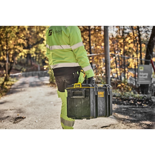 A person wearing high-visibility work clothing is carrying a DEWALT DWST83342-1 tool storage box outdoors on a sunny day, with trees in the background.