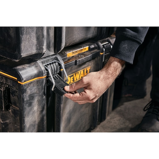 A close-up of a person’s hand unlocking the latch of a DEWALT ToughSystem 2.0 storage box, with visible DEWALT branding and rugged construction.