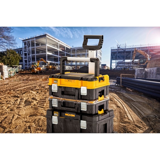 Stacked DEWALT TSTAK storage boxes with a telescopic handle shown on a construction site with building structures and construction equipment in the background.