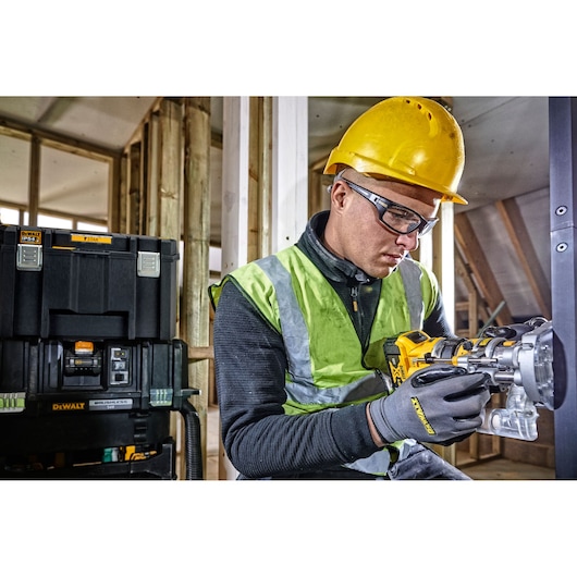 A worker wearing a yellow hard hat and high-visibility vest operates a power tool at a construction site. Next to the worker, there are DEWALT stackable tool storage boxes. The background shows unfinished wooden framing.