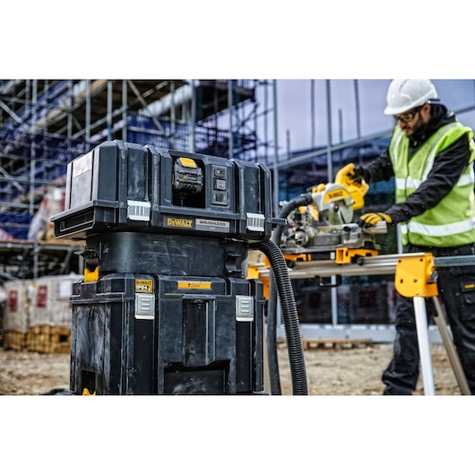 A DEWALT DWST83346-1 stackable toolbox and storage system is shown outdoors on a construction site, with a worker using a DEWALT circular saw in the background.