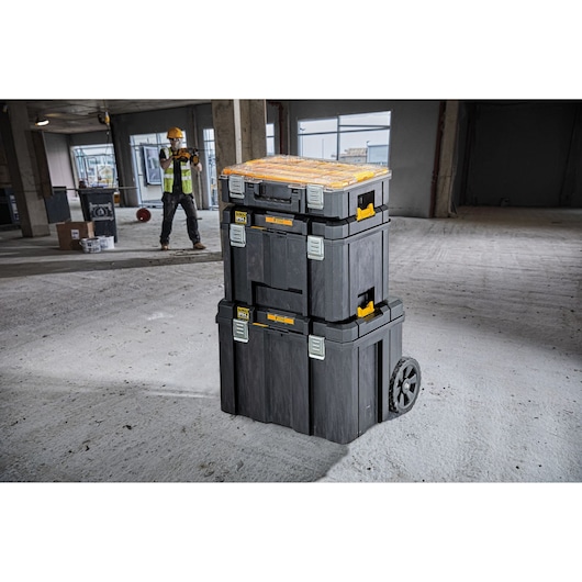 Stacked DEWALT toolboxes on wheels are shown in the foreground of a construction site with a worker in safety gear in the background.