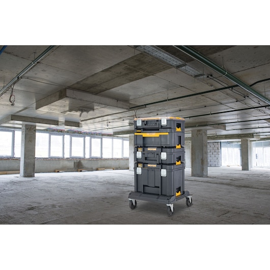 A stack of DEWALT black and yellow tool storage boxes mounted on a wheeled cart is positioned in the middle of an unfinished concrete construction site with large windows and exposed pipes.
