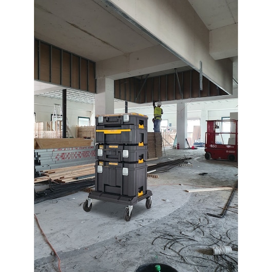 Stack of three DEWALT tool storage boxes on a wheeled cart in a construction site, with building materials and equipment visible in the background.