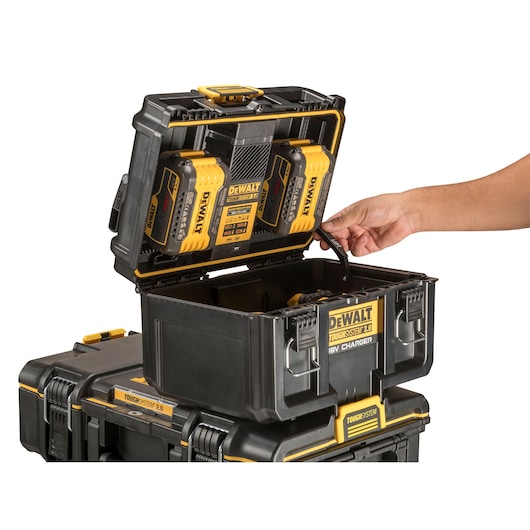 A DEWALT ToughSystem 2.0 storage box with an integrated 18V battery charger, shown open with two DEWALT batteries charging inside. A person's hand is holding a strap attached to the box.