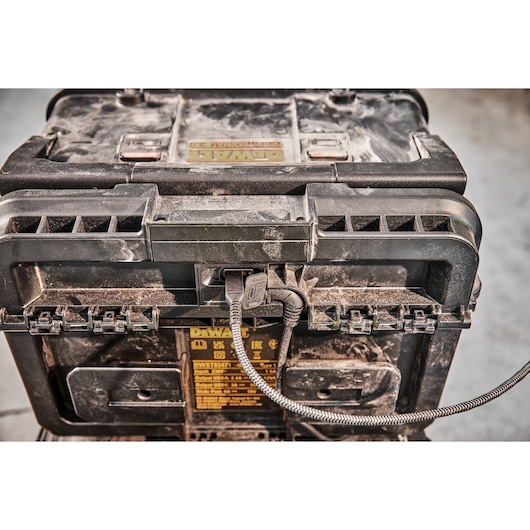 Close-up of a dusty DEWALT tough storage case with a charging cable plugged into its front panel. The case has visible branding and safety information sticker.