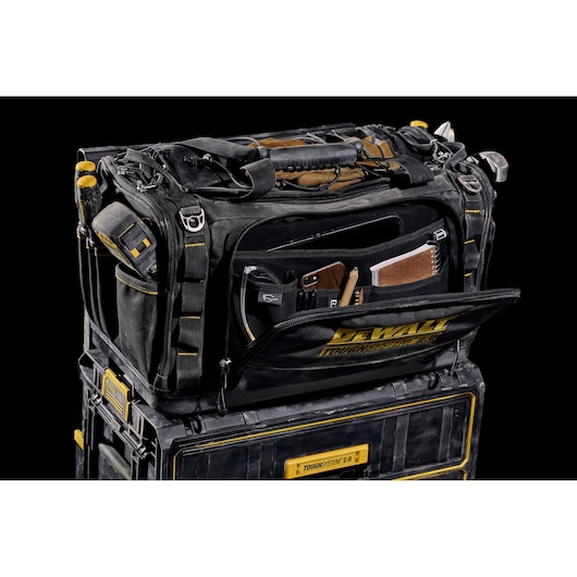 A black DEWALT tool bag with yellow accents, featuring multiple compartments and pockets, is shown opened and organized with various tools and accessories inside. The bag is placed on top of a DEWALT ToughSystem 2.0 storage box, both against a black background.