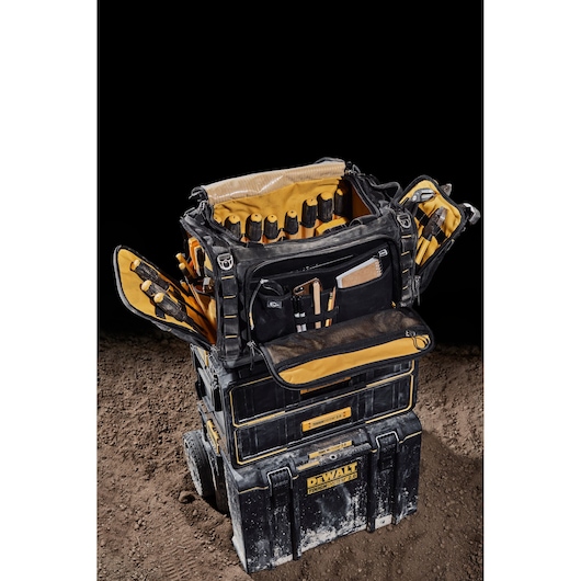 A DEWALT tool bag with multiple compartments and pockets, filled with various tools and accessories, is shown open and placed on top of two stacked DEWALT ToughSystem toolboxes on a dirt surface.