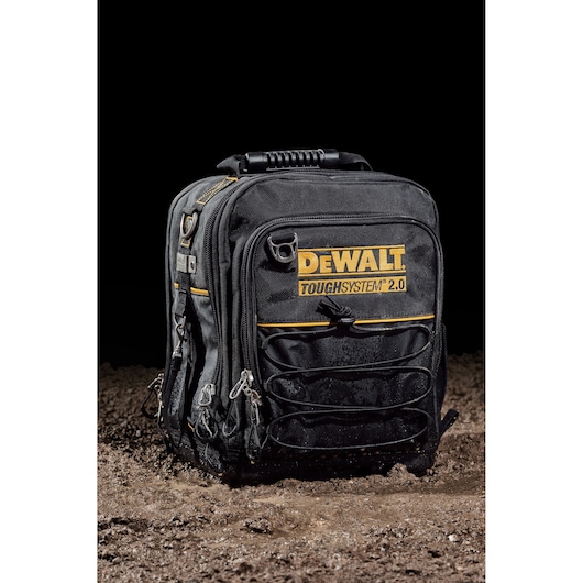 A black DEWALT ToughSystem 2.0 backpack with multiple zippered compartments and yellow accents, placed on dirt with a black background.