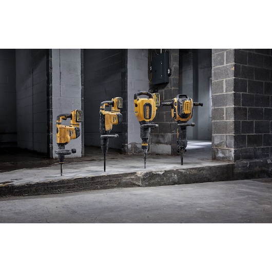 Four yellow and black DEWALT rotary hammer drills positioned upright with their drill bits on a concrete slab in an industrial setting with gray cinder block walls.