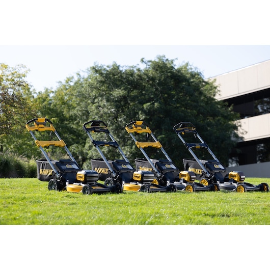 Four DEWALT cordless lawn mowers are lined up on a grassy lawn outside, with trees and a building in the background. The mowers are black and yellow and positioned at an angle, showing their side and front views.