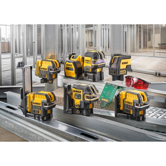 A group of DEWALT yellow and black laser level tools are displayed on metal construction framing, along with a pair of red safety glasses and a green measuring card. The background shows an unfinished building interior with exposed metal studs.