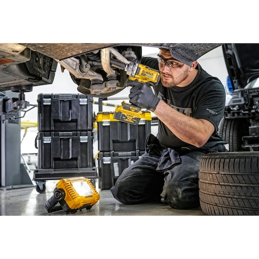 A mechanic uses a Dewalt cordless power tool to work on a car suspension. The scene includes several tool storage boxes, a portable work light, and a spare tire in a well-lit garage environment.