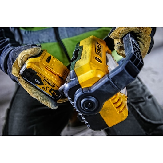 A person wearing work gloves is holding a yellow DeWalt power tool and attaching an 18V XR lithium-ion battery in an industrial or construction environment.