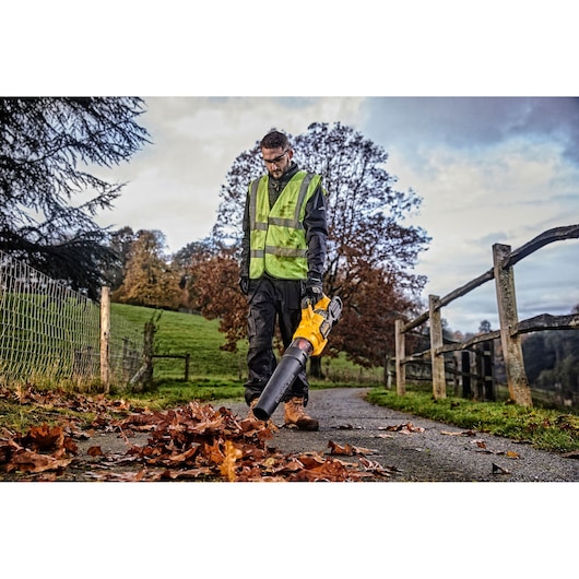 A person wearing a reflective safety vest is using a yellow leaf blower on a paved pathway covered with autumn leaves. The scene is outdoors, with trees, grass, and a wooden fence visible in the background.