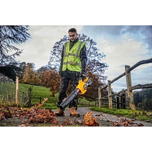 A person wearing a high-visibility vest and work boots is standing outdoors on a pathway with autumn leaves, holding a yellow and black leaf blower.