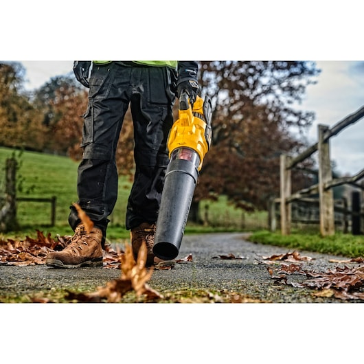 A person wearing work boots and black pants is using a yellow leaf blower to clear fallen leaves from a paved outdoor path. The surrounding area includes a wooden fence and grass, with trees in the background.