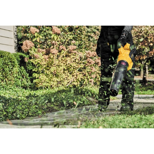 A person using a yellow and black cordless leaf blower to clear leaves and grass from a sidewalk in a garden area.