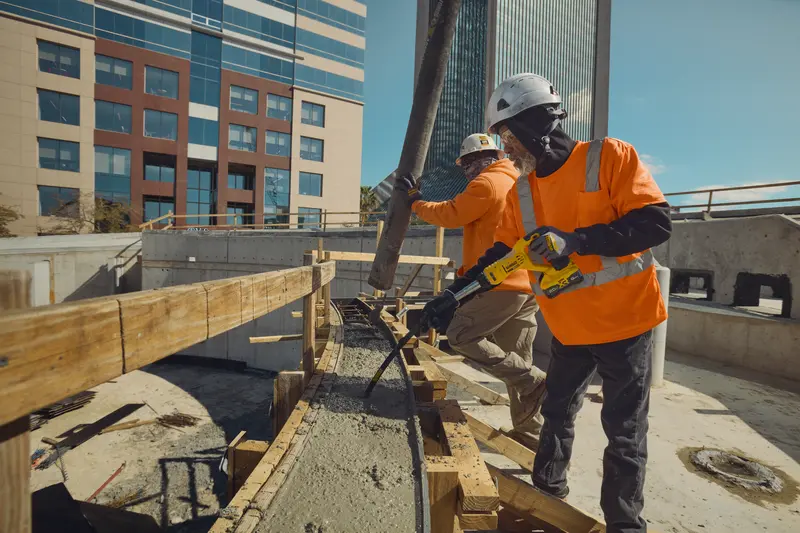 Two construction workers wearing safety helmets and orange high-visibility shirts are working at a construction site. One worker is using a DEWALT concrete vibrator tool on freshly poured concrete, while the other directs a concrete hose. Modern office buildings are visible in the background.