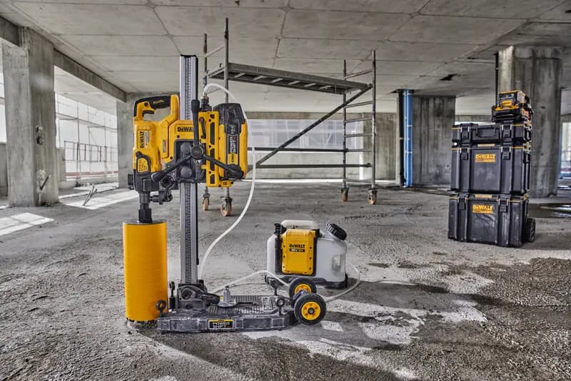 A DEWALT core drilling machine with a yellow cylindrical drill attachment and connected water tank is positioned on the floor of a concrete construction site. In the background, scaffolding and DEWALT branded stackable storage boxes are visible.