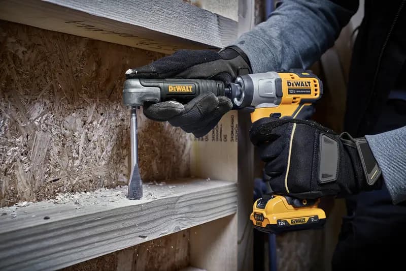 A person wearing gloves uses a DEWALT 12V cordless drill with a right angle attachment to drill into a wooden board, with wood shavings visible around the work area.