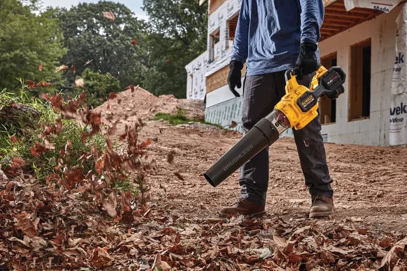 Person using a DEWALT cordless leaf blower to clear leaves on a dirt path near a construction site.