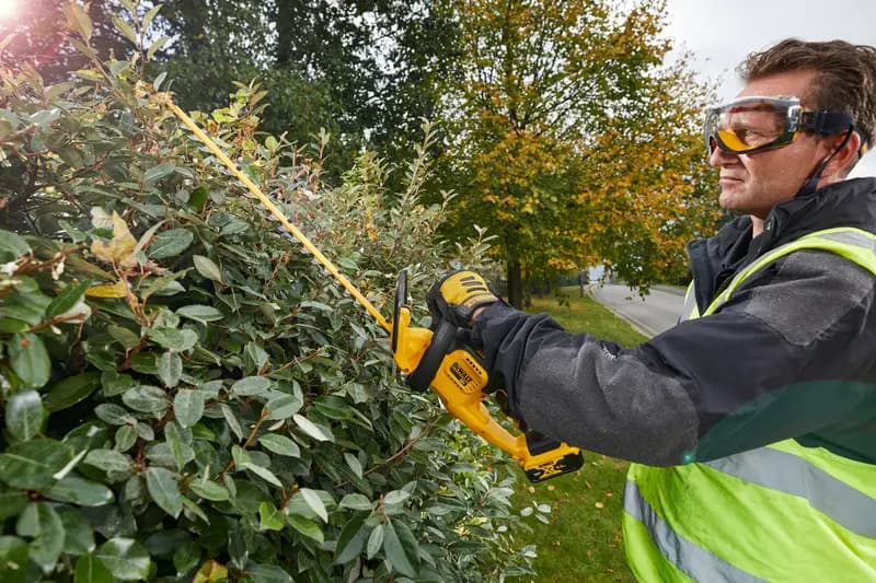 Photo of DEWALT hedge trimmer DCM563PB, DCM563PB-XJ used outdoors on bushes by worker in safety gear.