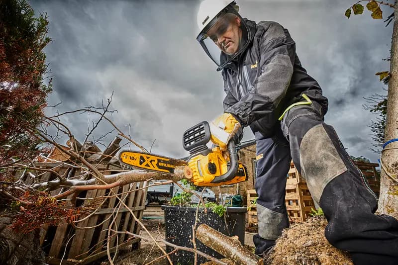 Photo of DEWALT DCM565N chainsaw cutting branches outdoors, worker in safety gear, yellow tool visible.