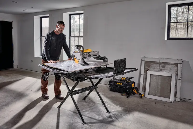 A person stands beside a DEWALT D36000S tile saw setup on its stand in a room under construction. The saw is placed on a foldable black stand, and there are tools and a DEWALT tool bag on the floor nearby. The room has unfinished walls, windows, and construction materials leaning against the wall.