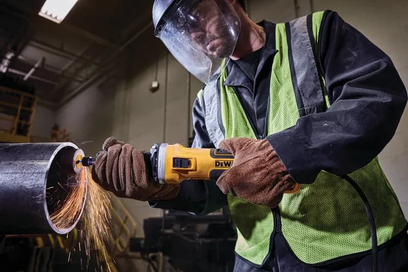 Worker using a DEWALT grinder on a metal pipe, wearing gloves, safety vest, and face shield with sparks flying.