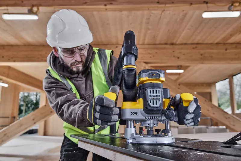 Worker uses DEWALT DCW620NT router on wood in a construction site, wearing gloves, vest and hard hat.