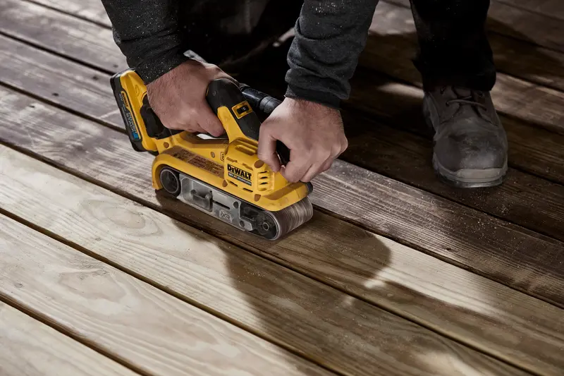 En person bruker en gul DEWALT trådløs båndsliper på treplanker for å jevne ut overflaten.