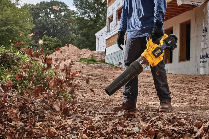 Person using a DEWALT cordless leaf blower to clear leaves on a dirt path near a construction site.