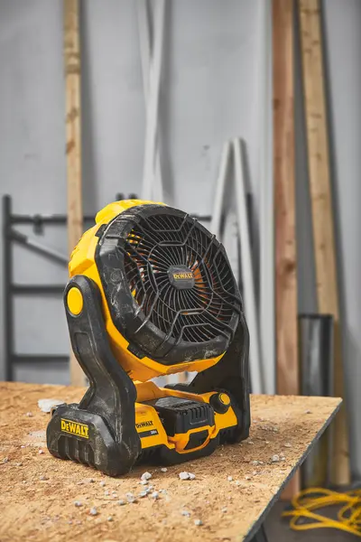 En bærbar, batteridrevet DEWALT-vifte (modell DCE512B) står på en arbeidsbenk av tre i et verksted med byggematerialer i bakgrunnen.