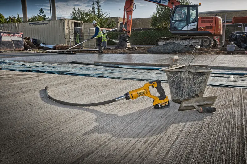 DEWALT cordless tool on a concrete site with bucket and trowel. Construction workers and excavator in background.